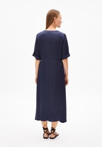 Mörkblå ljungklänning i knälängd tillverkad av slätt tyg, med rullade kortärmar och en rak bakseam. Ett par svarta platta sandaler visas.