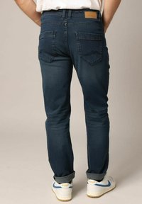 Jean en denim bleu foncé avec un design ajusté, poches arrière et une étiquette en cuir à la taille. Les ourlets retroussés révèlent des baskets blanches.