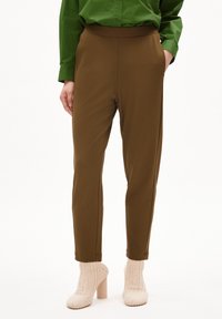 Brązowe zwężane spodnie wykonane z miękkiego materiału z wysokim stanem, w połączeniu z kremowymi dzianinowymi botkami do kostek. Zielona koszula z długimi rękawami.