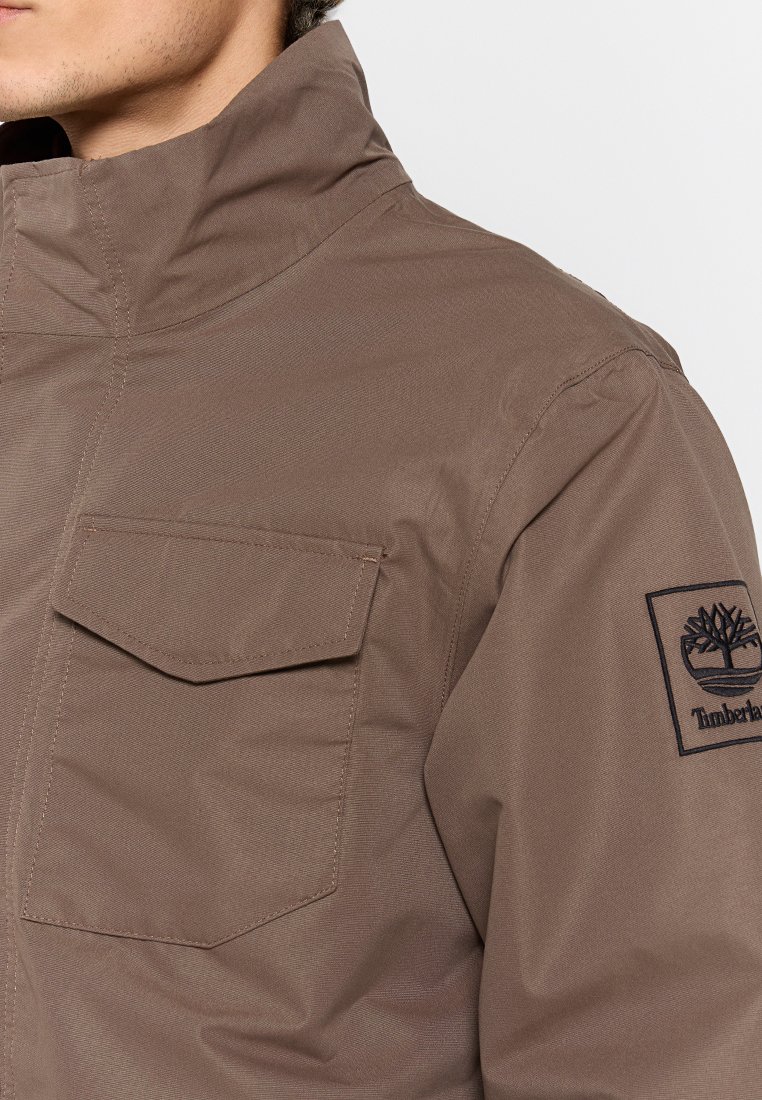 Primo piano di una giacca marrone con colletto alto, tasca sul petto con patta e patch del logo Timberland sulla manica superiore.