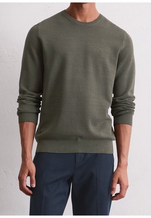Mann trägt einen strukturierten olivgrünen Langarmpullover mit gerippten Bündchen und Saum, kombiniert mit dunkelblauer Hose.