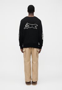 Μαύρο πλεκτό πουλόβερ με λευκό σχέδιο τρέχοντος σκύλου στην πλάτη, συνδυασμένο με μπεζ παντελόνι cargo και καφέ παπούτσια.