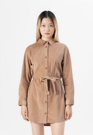 Vrouw met steil ombre haar, gekleed in een bruin corduroy hemdjurk met lange mouwen en een strikceintuur, staande tegen een witte achtergrond.