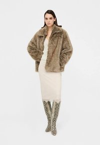 Bruine faux-fur jas, crèmekleurige gebreide top, nauwsluitende beige rok en kniehoogte slangenprintlaarzen. Zachte textuur, los ontwerp en gelaagde styling.