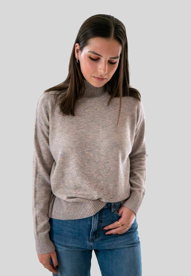 Jeune femme avec des cheveux bruns raides portant un pull à col roulé beige clair et un jean bleu, regardant vers le bas avec les mains près des poches.