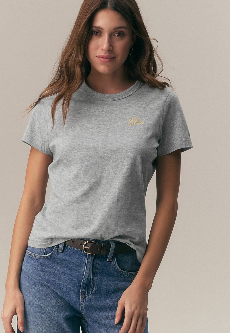 Siva bombažna majica z okroglim izrezom in kratkimi rokavi, s subtilnim rumenim vezenjem na prsih, v kombinaciji s temnimi kavbojkami iz jeansa.