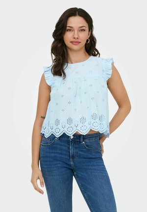 Moteris dėvi šviesiai mėlyną be rankovių viršutinę dalį su akutėmis, raukiniais ant pečių ir banguotu kraštu, derintą prie mėlynų džinsų, stovinčią prieš paprastą foną.