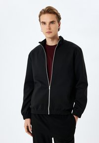 Veste zippée noire en tissu lisse. Présente un col haut, des poignets et une taille élastiques. Comprend une fermeture éclair argentée et des poches latérales.