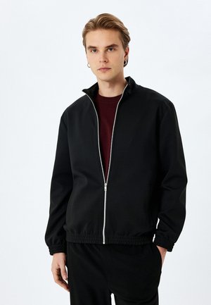 Chaqueta negra con cremallera fabricada en un tejido suave. Cuenta con un collar alto, puños y cinturilla elásticos. Incluye una cremallera plateada y bolsillos laterales.