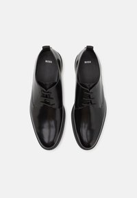 Chaussures habillées noires en cuir avec une finition lisse, dotées d'un design à lacets, d'un bout rond et d'une semelle texturée. Logo visible sur la semelle intérieure.