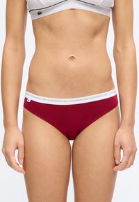 Γυναίκα που φοράει κόκκινα εσώρουχα Lacoste και άσπρο αθλητικό σουτιέν, εμφανιζόμενη από τη μέση έως τους πάνω μηρούς σε απλό φόντο.