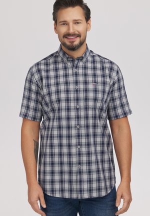 Mężczyzna uśmiechający się, ubrany w szarą i granatową koszulę w kratę z krótkim rękawem na guziki, z widocznym tatuażem na lewym przedramieniu, stojący na jednolitym tle.