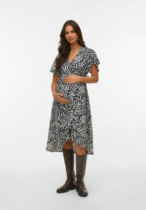 Femme enceinte portant une robe à motifs bleu marine et blanc et des bottes marron jusqu'aux genoux, tenant doucement son ventre, debout sur un fond blanc.
