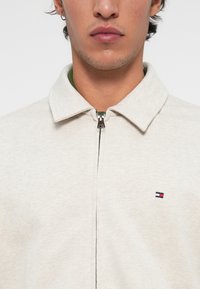 Jasnoszara kurtka zapinana na zamek z wyprofilowanym kołnierzem, z zieloną podszewką i małym logo po lewej stronie klatki piersiowej. Gładka tekstura.