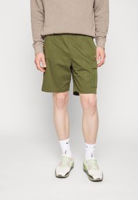Grüne Cargo-Shorts mit einem elastischen Bund und einer Seitentasche, kombiniert mit weißen und grünen Sportschuhen und weißen Socken.