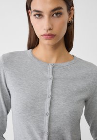 Kvinna med rakt brunt hår som bär en ljusgrå ribbad knapplös cardigan, vänd framåt med en neutral uttryck.