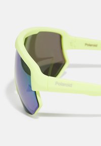 Sárgászöld Polaroid sport napszemüveg, körbeölelő sötét, tükröződő lencsékkel és vastag szárakkal, fehér háttéren.