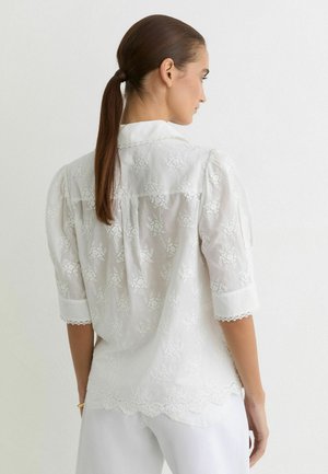 Barna lófarokkal rendelkező nő fehér virágos csipkeblúzt visel, hullámos szegéllyel és félhüvelyű ujjakkal, sima fehér háttér előtt áll.