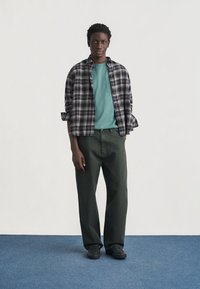 Νεαρός άνδρας που στέκεται φορώντας μπλουζάκι τιρκουάζ, μακρυμάνικο πουκάμισο σε ασπρόμαυρο καρό, σκούρο πράσινο παντελόνι και μαύρα παπούτσια σε μπλε πάτωμα.