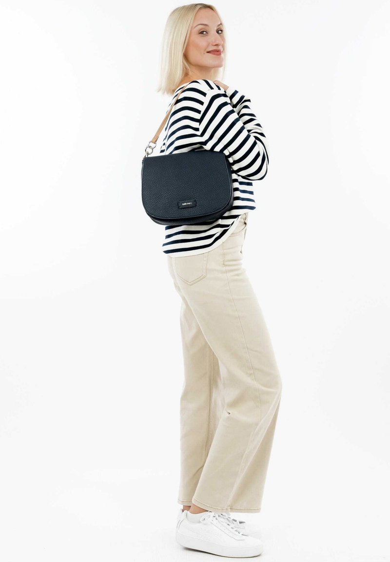 Navy blue textured leather crossbody bag with a curved shape and a silver hardware accent. Model wears striped top and cream-colored pants.