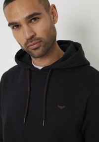 Gros plan sur un homme aux cheveux courts et foncés avec une barbe, portant un hoodie noir avec un petit logo rouge brodé près de la poitrine.