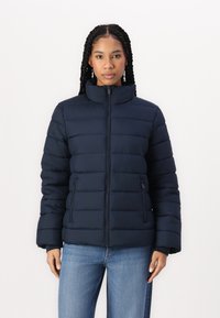Geacă puffer bleu închis cu guler înalt, cusături orizontale, buzunare cu fermoar și croială lejeră. Textură netedă, design izolat.