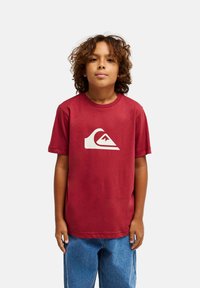 Κόκκινο βαμβακερό t-shirt με κοντά μανίκια, με μεγάλο λευκό λογότυπο κύματος στο μπροστινό μέρος. Φορεμένο με μπλε τζιν τζιν.