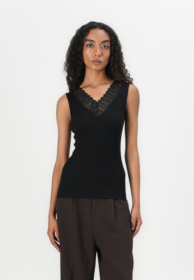 Canotta nera a coste con un bordo in pizzo decorativo lungo il collo a V. Presenta una silhouette aderente e design senza maniche, abbinata a pantaloni marroni.