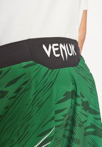 Zöld rövidnadrág merész fekete grafikai mintákkal, fekete derékrésszel, amelyen fehér "VENUM" logó található, sima anyagból készült, lazább szabással.