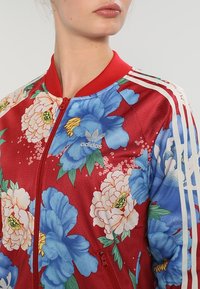 Röd blommig bomberjacka med stora blå och vita blommor, blommönster och tre vita ränder längs ärmarna; dragkedja.
