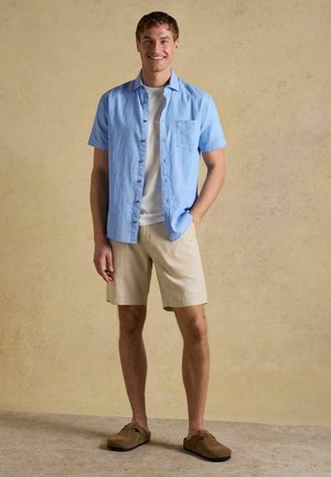 Junger Mann steht, trägt ein hellblaues kurzärmeliges Hemd über einem weißen T-Shirt, beige Shorts und braune Slip-on-Schuhe und lächelt.