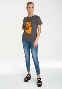 Graues T-Shirt mit einem Cartoon-Charakter mit orangefarbenem Körper und braunen Gliedmaßen, getragen mit blauen Jeans und schwarzen Plateauschuhen.