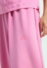 Svetloružové teplákové nohavice vyrobené z mäkkého materiálu, ktoré majú embosované logo Adidas na ľavom stehne a bočné vrecká na uloženie.