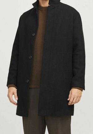 Uomo che indossa un cappotto nero con bottoni sopra un maglione marrone e pantaloni marroni, in piedi contro uno sfondo chiaro e semplice.