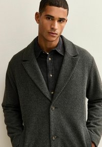 Cappotto di lana grigio scuro con colletto a scialle, chiusura con un solo bottone e due tasche laterali, indossato sopra una camicia nera a bottoni con texture.