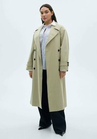 Cappotto impermeabile beige con design doppiopetto, bottoni neri e grandi revers. Abbinato a una camicia azzurra chiara e pantaloni scuri.