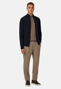 Jūrininkų zip-up džemperis virš tan polo marškinėlių, derinamas su šviesiai smėlinėmis kelnėmis ir tamsiais sportbačiais. Yra modernus dizainas ir glausta išvaizda.