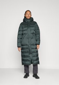 Duga tamnozelena puffer jakna s visokim ovratnikom, prednjim zatvaračem i velikim džepovima. Izrađena od sjajnog, kviltanog materijala s glatkom teksturom.
