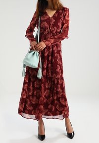 Robe longue bordeaux avec un motif floral, manches transparentes et design à étages. Associée à un sac à main vert menthe avec des accents de pompons.