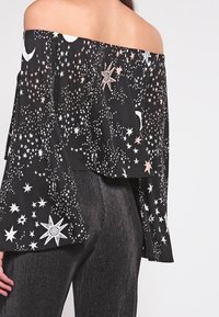 Blusa negra sin mangas con mangas acampanadas, con un patrón de estrellas y lunas en blanco y acentos en oro rosa. Tejido texturizado.
