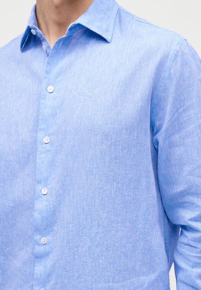 Ανοιχτό μπλε πουκάμισο με μακριά μανίκια και κουμπιά, με γιακά και διακριτικό κεντημένο λογότυπο στο αριστερό στήθος, φορεμένο από ένα άτομο.