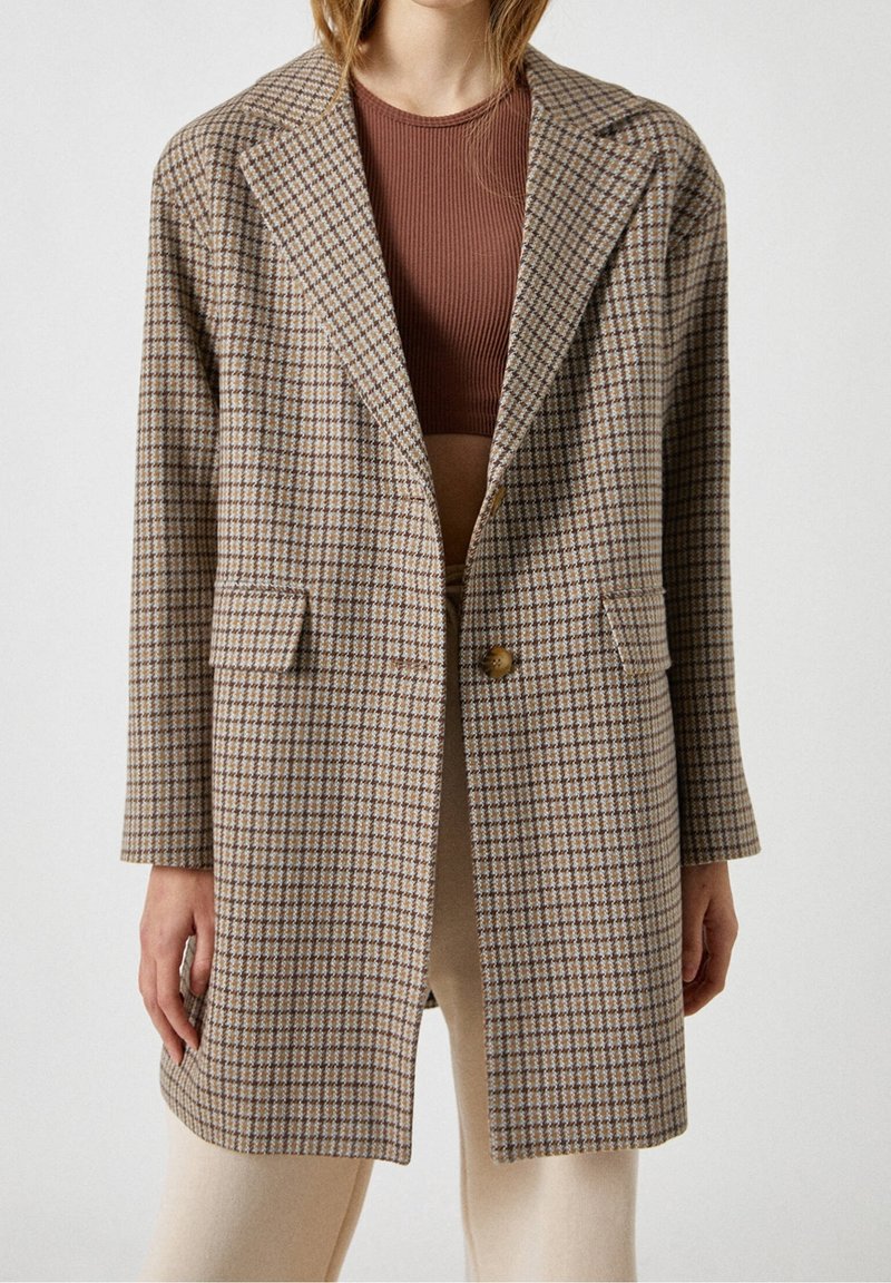 Personne portant un long blazer marron à motifs par-dessus un top court côtelé marron et un pantalon beige clair, debout devant un fond uni.