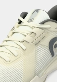 Λευκό αθλητικό παπούτσι με υφασμάτινο επάνω μέρος με υφή, ενσωματωμένο σύστημα κορδονιών και γκρι λεπτομέρειες. Διαθέτει σήμανση "Deltastrap" κοντά στα κορδόνια.