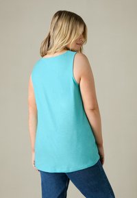 Un top senza maniche di un leggero color turchese, realizzato in un tessuto morbido, con scollatura rotonda e orlo leggermente curvo, indossato con jeans scuri.