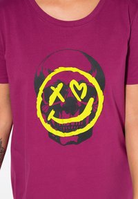 Magenta T-shirt lavet af bomuld med en sort kraniets grafik, der har et gult smilende ansigt og hjerteformede øjne. Rund halsudskæring, korte ærmer.