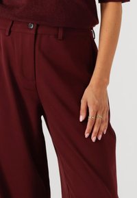 Bordeaux op maat gemaakte broek met een zachte textuur, voorzien van een voorzak en een knoopsluiting. Een hand versierd met gouden ringen rust aan de zijkant.