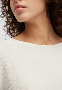 Jasno beżowy, dziany top z szerokim dekoltem, z teksturowaną tkaniną i subtelnymi detalami szwów wokół kołnierza i ramion.
