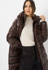 Brun pufferjakke med en blank overflade, udstyret med hætte, lynlås og vandret quiltet mønster. Båret over en ribbet cremefarvet sweater.