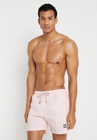 Costumi da bagno rosa chiaro realizzati in tessuto leggero, con vita elastica e cordoncino di regolazione, e una piccola targhetta con logo sull'orlo.