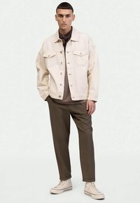 Mladý muž oblečený v béžovej bunde, hnedých nohaviciach, tmavom tričku a béžových vysokých teniskách, stojí na jednolatom svetlom pozadí.